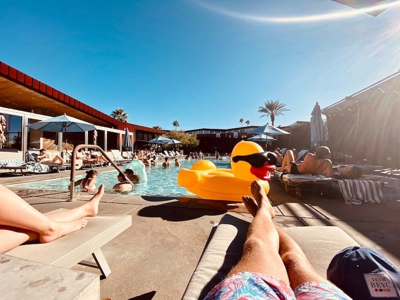 Swimmers lounging in and around a pool in sunny Palm Springs with a large blow up duck floatie with sunglasses.