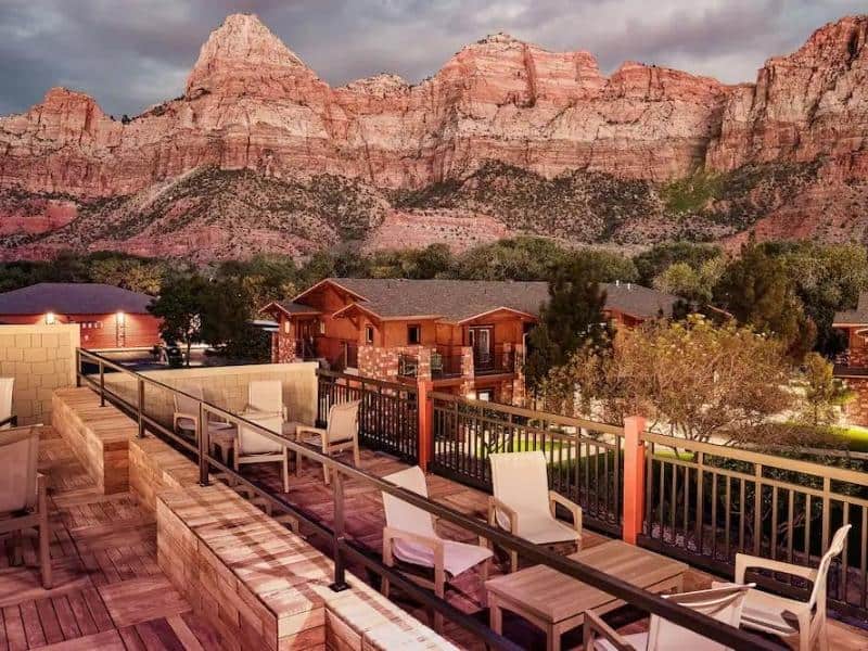 Outdoor balcony with chairs and tables overlooks Cable Mountain Lodge with red rocks in the background near Zion National Park