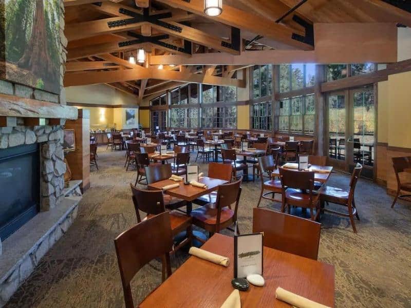 Spacious dining hall at John Muir Lodge with stone fireplace and large windows offering views of the surrounding forest. Sequoia and Kings Canyon National Park lodging option.