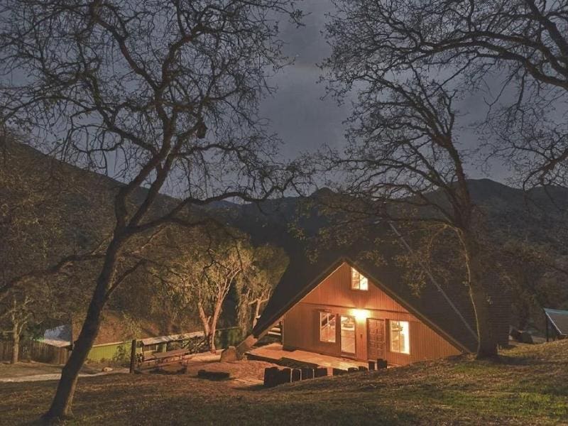 A-frame cabin illuminated at twilight with bare trees in the foreground. A Vrbo located inside the Sequoia National Park gates. Where to stay in Sequoia National Park.