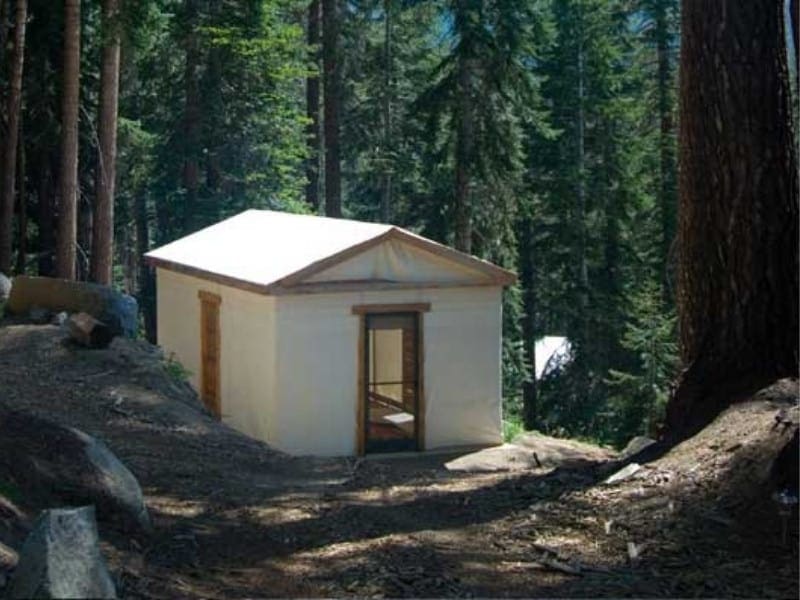 Simple white cabin with a wooden door, nestled among tall pines at Sequoia High Sierra Camp, a hike-in glamping property in Sequoia National Park.