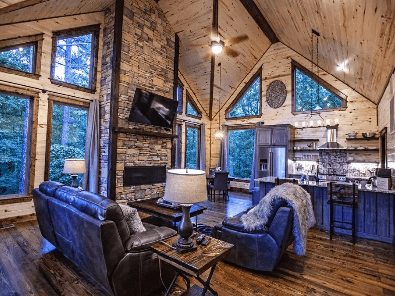 Interior view of a Blue Magnolia luxury cabin in Broken Bow featuring a living room with blue leather sofas, a stone fireplace, and a mounted TV against a backdrop of forest views through large windows.
