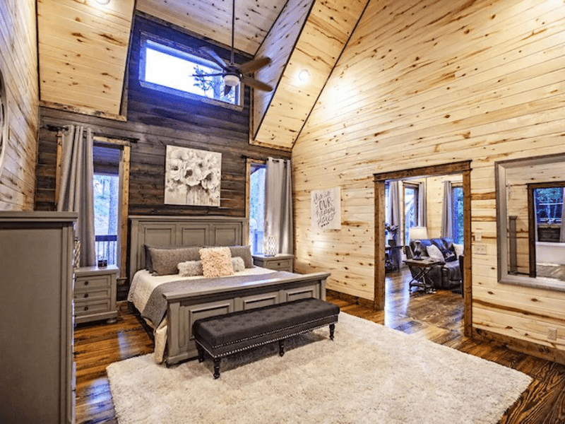 Bedroom inside a Broken Bow luxury cabin rental, with a large bed, overhead loft window, rustic wooden walls, and artwork, leading to an adjoining living space.