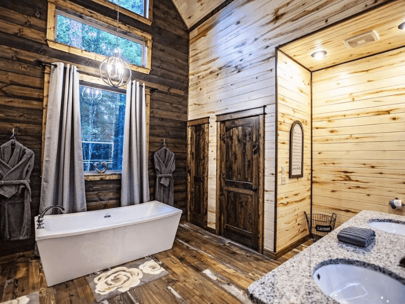 Bathroom of a Broken Bow Vrbo rental with a freestanding bathtub, wooden cabinets, and plush robes hanging next to a window with forest views.