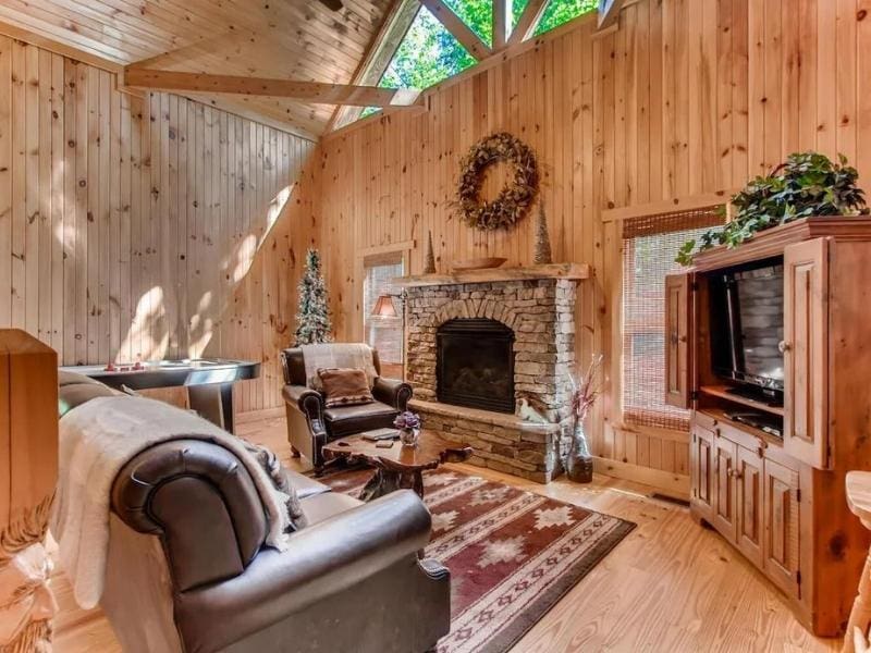 Rustic cabin living room with vaulted ceilings and ample natural light from skylight windows. The room has a stone fireplace, comfortable leather seating, and cabin-style decor including wildlife-themed artwork.