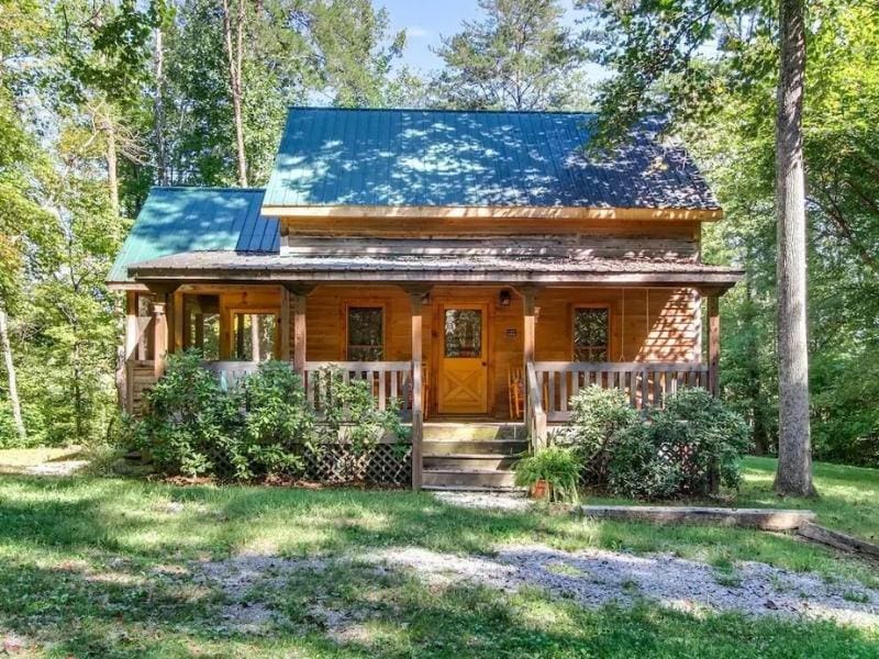 Exterior view of a traditional log cabin in Pigeon Forge surrounded by greenery in a forest setting. The cabin features a quaint front porch.