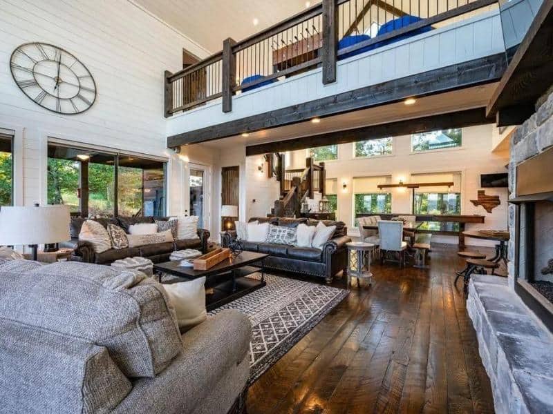 Open-plan living space of 'High Faultin' Hideaway' with a large wall clock, plush seating, and a loft-style bedroom overhead. Luxury cabin in Broken Bow.
