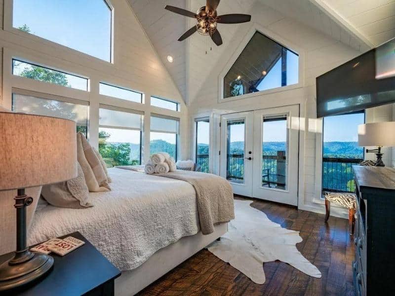 Bedroom in 'High Faultin' Hideaway' in Broken Bow featuring vaulted ceilings, large windows with forest views, and French doors leading to a balcony.