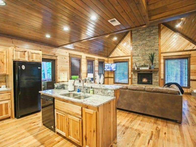 Open-concept kitchen with a granite island and stainless steel appliances, flanked by a cozy living area with a fireplace in a Broken Bow cabin rental.