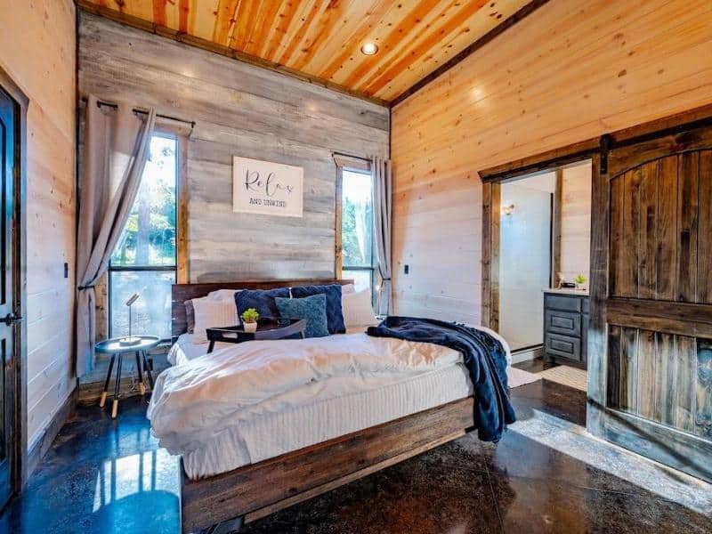 Bedroom in 'Somethin 2 Talk About' Broken Bow cabin with wooden paneling, a plush white bed, and 'Relax and Unwind' wall art.