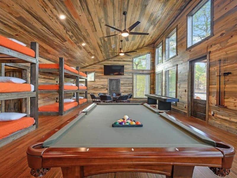 Game and bunk room in 'StarFire' luxury Broken Bow cabin featuring multiple bunk beds with orange bedding, a pool table, and large windows offering forest views.