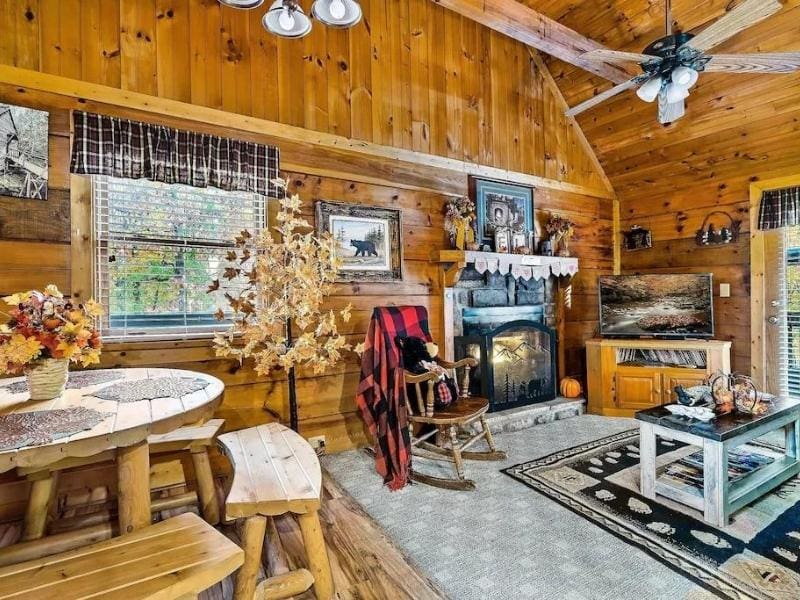 Rustic dining and living area in a cabin rental in Pigeon Forge with natural wood interiors and large windows providing ample natural light. The space features a unique round wooden dining table, cozy seating areas, and a stone fireplace with festive autumn decorations and wildlife art.