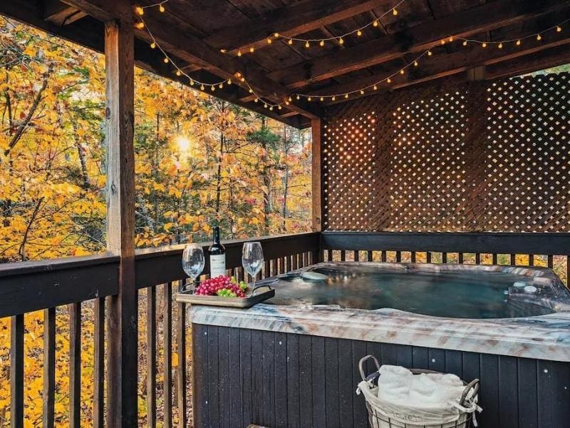 Outdoor hot tub on a covered deck with string lights, overlooking a stunning autumn forest scene. The setting includes a wine bottle and glasses.