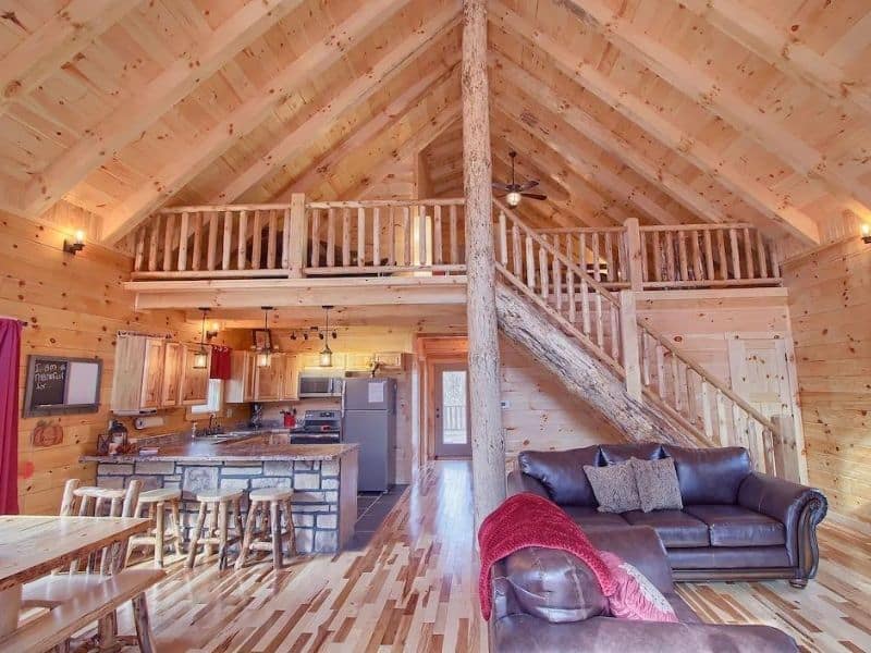 Open-plan interior of a Hocking Hills cabin featuring a high vaulted ceiling with exposed beams, a central wooden column, and a loft area above the kitchen.