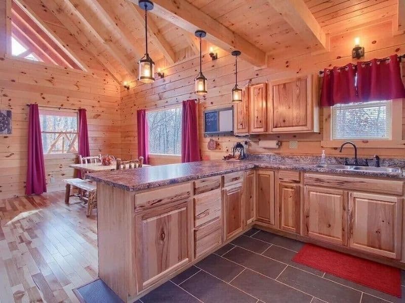 Rustic kitchen in a cabin with natural wood cabinets, granite countertops, and red accents from window curtains.