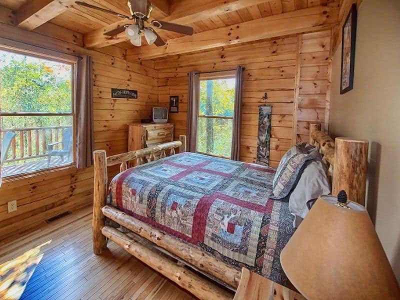 Cozy log cabin bedroom with a rustic quilted bed, wooden walls, and large windows offering views of the surrounding forest at Liberty Ridge Pet-Friendly Cabin in Hocking Hills, Ohio.