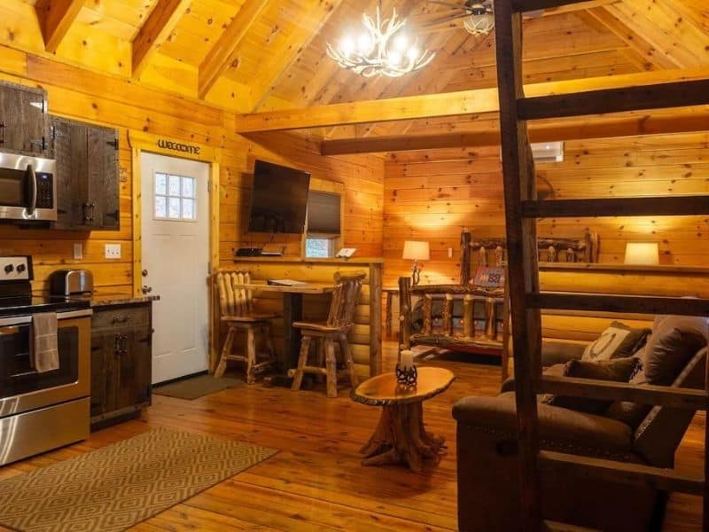 A cozy cabin interior with wooden walls and vaulted ceilings. The space includes a compact kitchen with stainless steel appliances, a small dining area with wooden stools, and a seating area. A ladder leads up to a loft space. The Peak @ Pine Creek in Hocking Hills, OH.