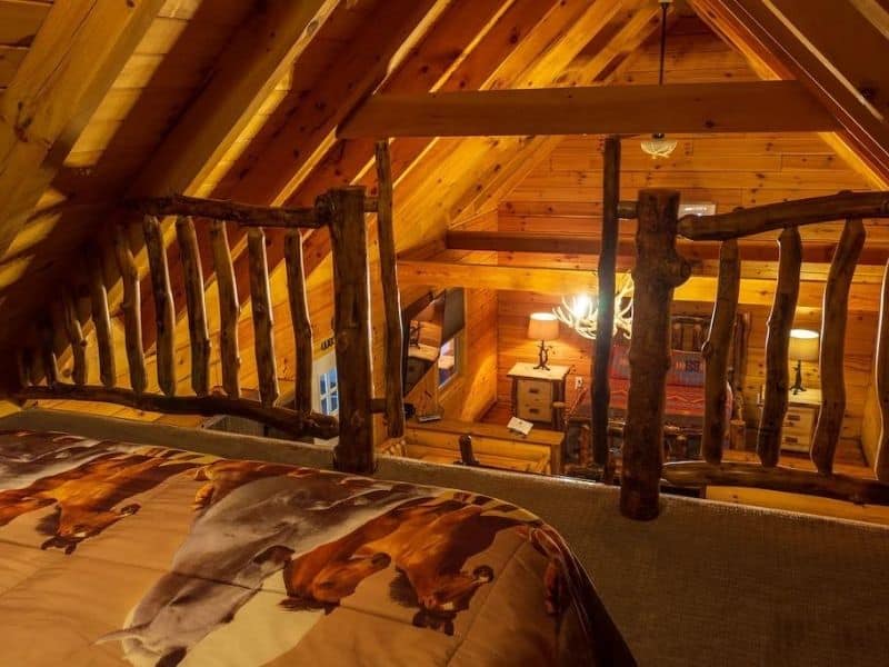 The Peak Cabin @ Pine Creek in Hocking Hills, OH. A view from the loft space overlooking the main living area below. The loft features wooden railings and cozy bedding with nature-themed decor.