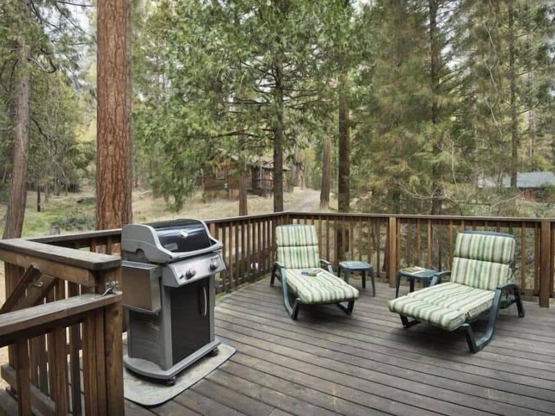 Spacious wooden deck of a Yosemite cabin rental equipped with a barbecue grill and outdoor recliners, set amidst pine trees.