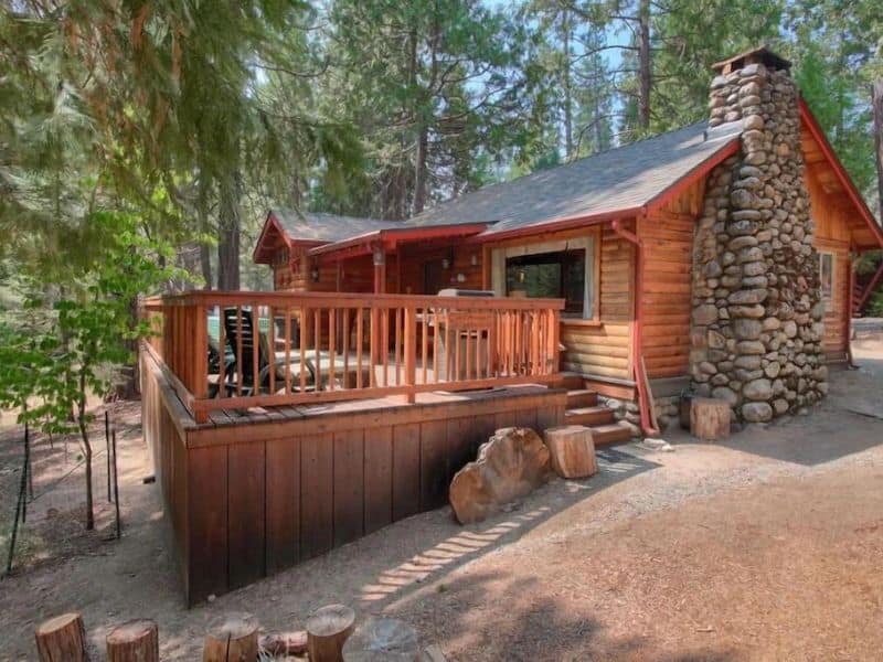 Rustic log cabin in Yosemite with a stone chimney and wooden front deck surrounded by forest trees.