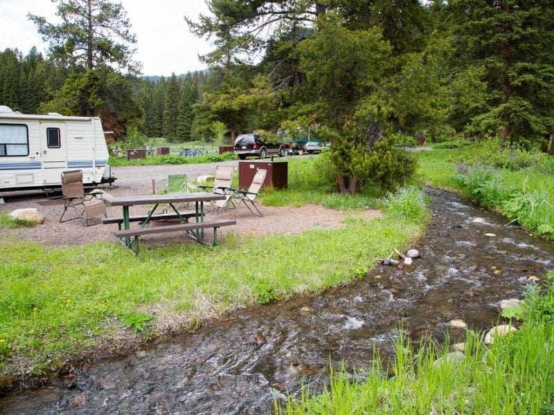 A campsite located right next to a running stream with a picnic table and fire ring at Pebble Creek Campground in Yellowstone National Park.