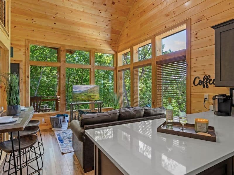 Living area of Pigeon Forge cottage rental with tall windows with forest view, kitchen, seating area, and dining table in a single room cabin, with colorful details.