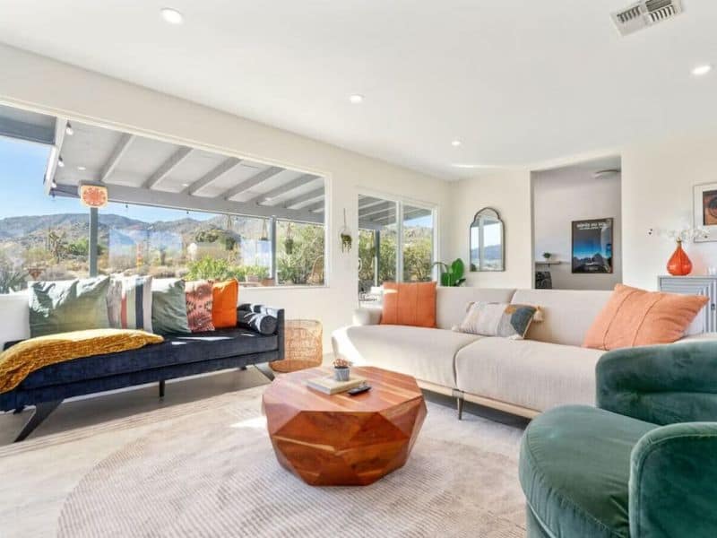 A bright living area with large windows offering panoramic desert views. The room features a modern sectional sofa with colorful throw pillows and a stylish geometric wooden coffee table. Pueblo Trail Vrbo in Joshua Tree.