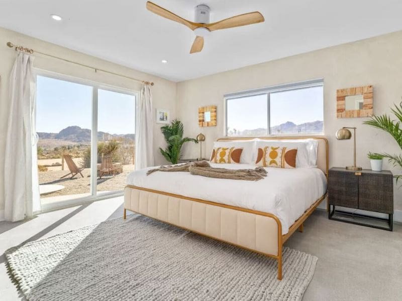 A bedroom in a Joshua Tree rental with a large bed dressed in white linens and accented with orange and yellow throw pillows. The room has a ceiling fan, light-colored curtains, and large sliding glass doors that open to an outdoor seating area with desert views.
