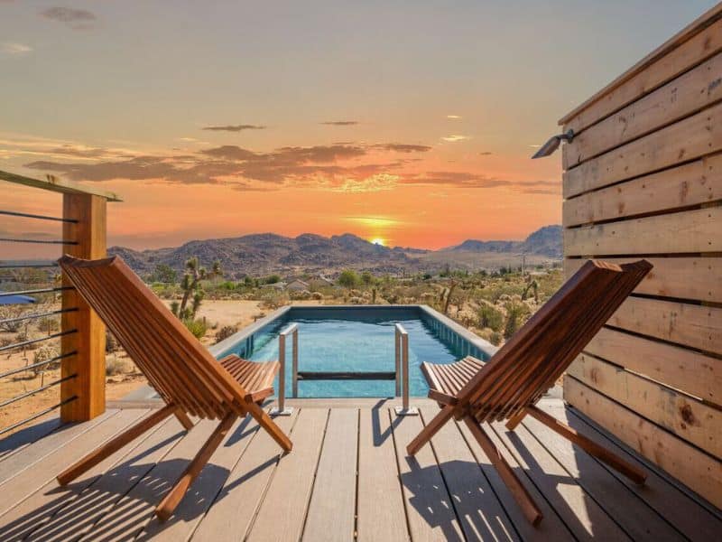 A picturesque outdoor pool area with a view of the desert at sunset. Two wooden lounge chairs are on a deck overlooking the pool. The pool is surrounded by desert vegetation. One of the prettiest vacation rentals near Joshua Tree.