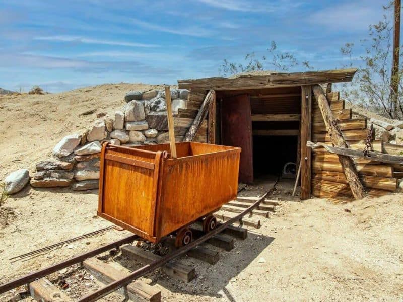 A rustic, wooden entrance to an old mining tunnel with an antique mining cart on tracks in the foreground. The entrance is surrounded by a rocky desert landscape.