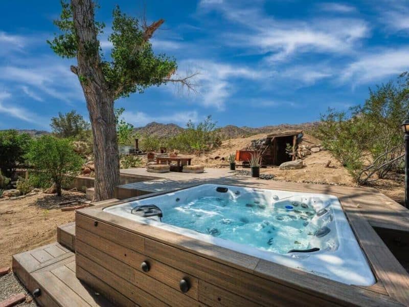 An outdoor hot tub set on a wooden deck, surrounded by natural desert scenery. An old mining cart and tunnel are visible in the background, adding to the unique character of the property. @ Marbella Lane - Vrbo in Joshua Tree.