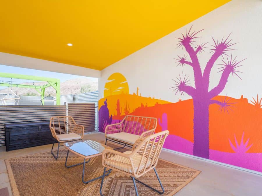 A vibrant outdoor seating area with a bright yellow ceiling and a colorful mural of a desert landscape featuring cacti and a sunset. The space includes wicker furniture with a glass-top table and patterned outdoor rugs. Best Airbnbs in Joshua Tree for Couples.
