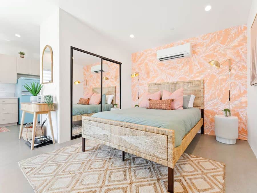 A stylish bedroom with a woven bed frame, green bedding, and pink accent pillows. The room has a peach-colored feature wall with a white leaf pattern, gold wall lamps, and a large mirrored closet door. There is also a wooden console table with a plant and decorative items. Part of a guide to the prettiest Airbnbs and Vrbos in Joshua Tree.