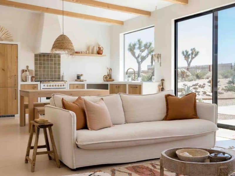 A bright and airy living area with a comfortable cream-colored sofa with earthy-toned throw pillows. The open-plan space includes a modern kitchen with wooden cabinets, a dining area, and large windows that offer views of the Joshua Tree desert. Natural light floods the room, highlighting minimalist, bohemian decor.