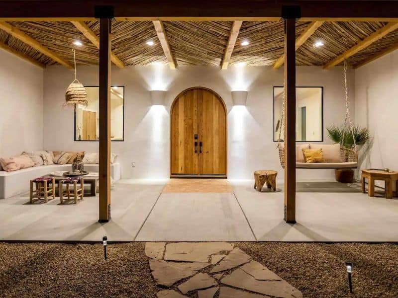 The front entrance to Desert Wild, featuring a wooden arched door under a rustic, thatched roof. The porch area is lit with soft lighting, and includes comfortable seating with a mix of cushions and throw pillows. A hanging swing adds a charming touch to the welcoming entryway. popular Airbnb rental in Joshua Tree, CA.