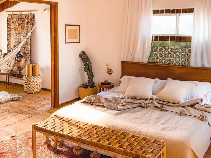 A cozy bedroom with a wooden bed with white linens and decorative pillows. The room has a warm, earthy ambiance with terracotta tile flooring, woven accents, and a large cactus in the corner. A woven bench sits at the foot of the bed, and an adjoining space with a hammock adds a bohemian touch. One of the most popular Airbnbs in Joshua Tree.