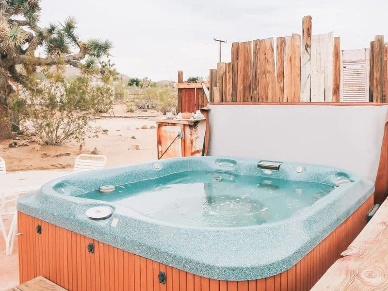 An outdoor hot tub in a rustic, fenced area in Joshua Tree, offering a relaxing spot to unwind and enjoy the desert landscape. The hot tub is surrounded by natural wooden fencing and desert plants, providing privacy and a serene atmosphere for guests.