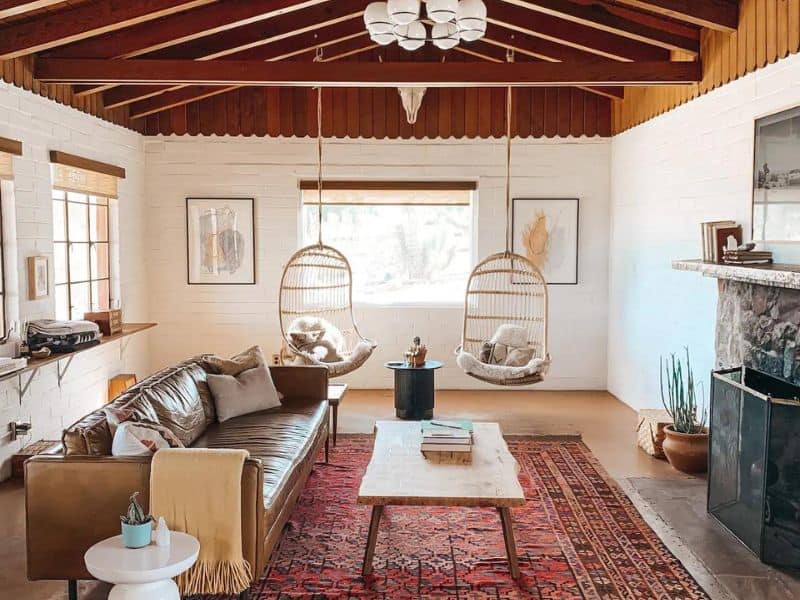 A charming living room with a rustic feel, featuring a leather sofa, a wooden coffee table, and a patterned red rug. The room includes unique hanging wicker chairs, exposed wooden beams on the ceiling, and a stone fireplace, creating a cozy and inviting atmosphere. One of the prettiest Joshua Tree Airbnbs.