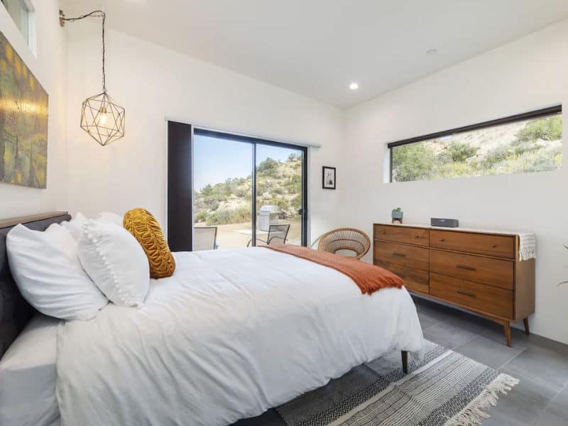 A cozy bedroom with a bed dressed in white linens and accented with orange and mustard pillows. The room has a mid-century modern dresser, a woven chair, and large windows that provide natural light and views of the surrounding desert landscape. Part of a guide to the Prettiest Joshua Tree Airbnbs and Vrbos.