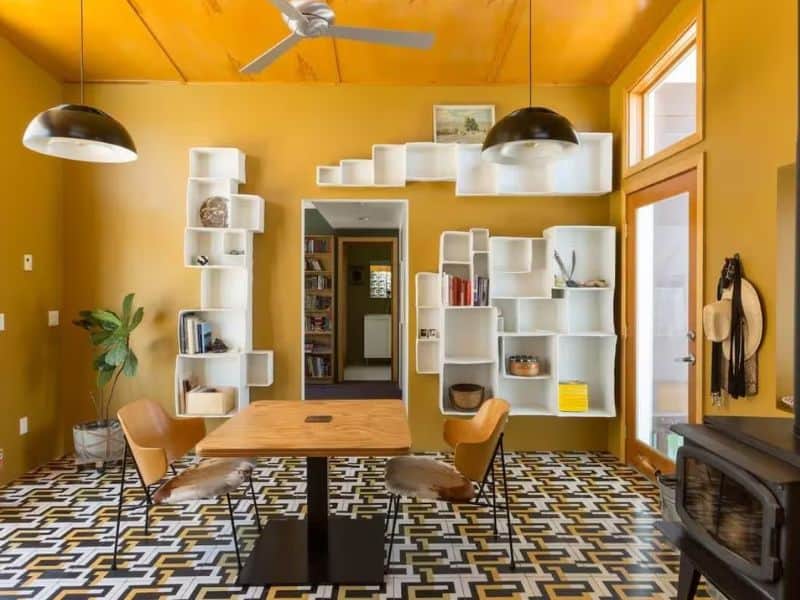 A stylish dining area featuring bold mustard-yellow walls and unique black and white patterned tile flooring. The room includes a square wooden table with two modern chairs and eclectic white shelving units displaying various decorative items. Two large pendant lights hang from the ceiling. Vacation rental in Joshua Tree, CA.