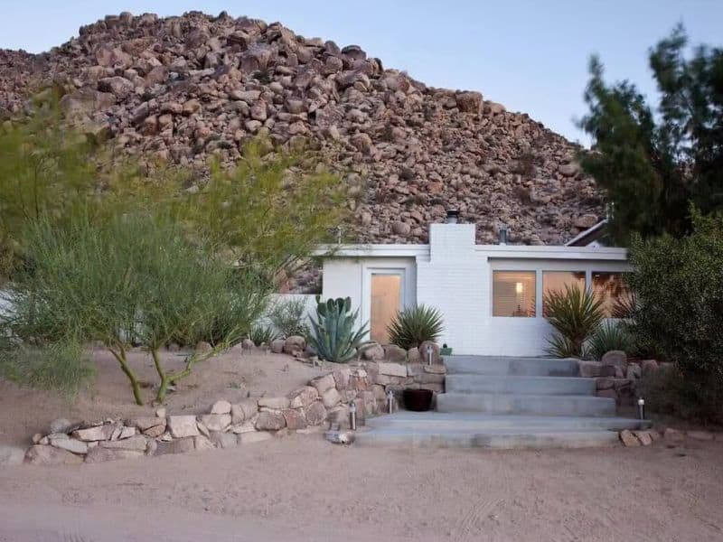 The exterior of Andrea Zittel's A Z West House in Joshua Tree, set against a rugged rocky hillside. The white brick house is surrounded by desert plants and features a simple, clean design with large windows that offer a glimpse of the interior. A set of concrete steps leads up to the front entrance, blending with the natural landscape.