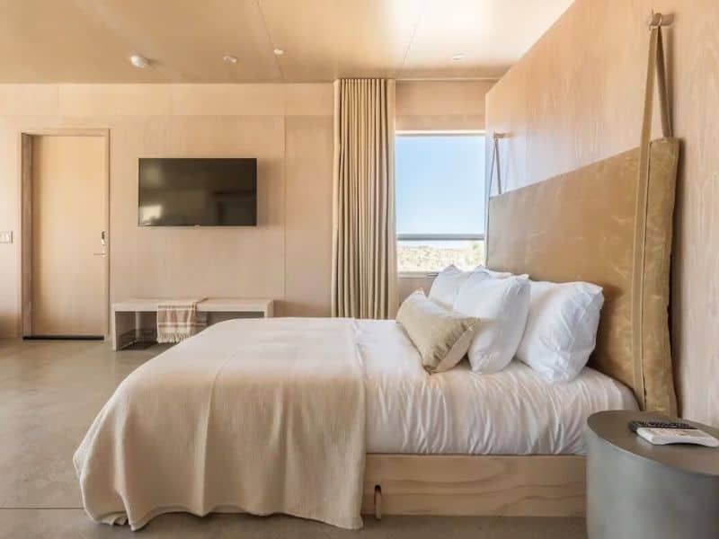 A spacious bedroom with a calming, neutral color palette. The room includes a large bed with crisp white linens and a light wooden headboard. A wall-mounted flat-screen TV is on one side, and natural light streams in from a window with beige curtains.