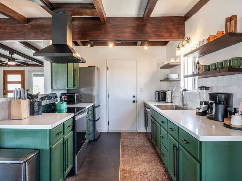 A modern kitchen with a blend of rustic and contemporary design elements. The kitchen features dark green cabinets, white countertops, and stainless steel appliances. Exposed wooden beams on the ceiling and open shelves add a touch of rustic charm. A decorative rug runs along the length of the kitchen.