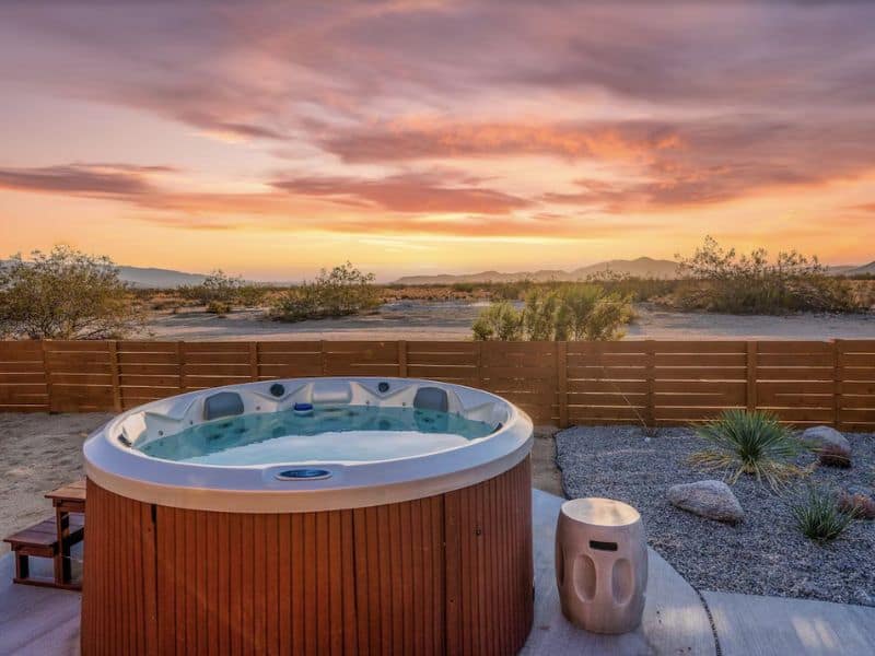 An outdoor hot tub set against a desert sunset backdrop. The circular hot tub is surrounded by a concrete deck and a gravel yard with desert plants. A wooden fence encloses the area. One of the best vacation rentals in Joshua Tree.