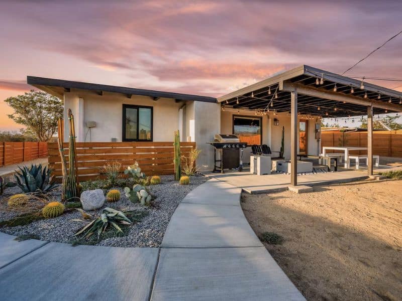 The exterior view of JT Haus with a covered patio area with string lights, a barbecue grill, and outdoor seating. The patio is adjacent to a landscaped yard with various desert plants. The sky is painted with the warm hues of a desert sunset. Part of a guide to the best Airbnbs and Vrbos in Joshua Tree.