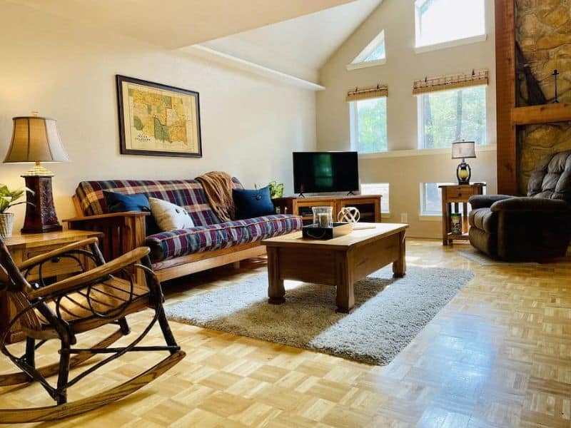 Living room with a plaid couch, wooden coffee table, TV on a stand, rocking chair, and large windows allowing natural light.
