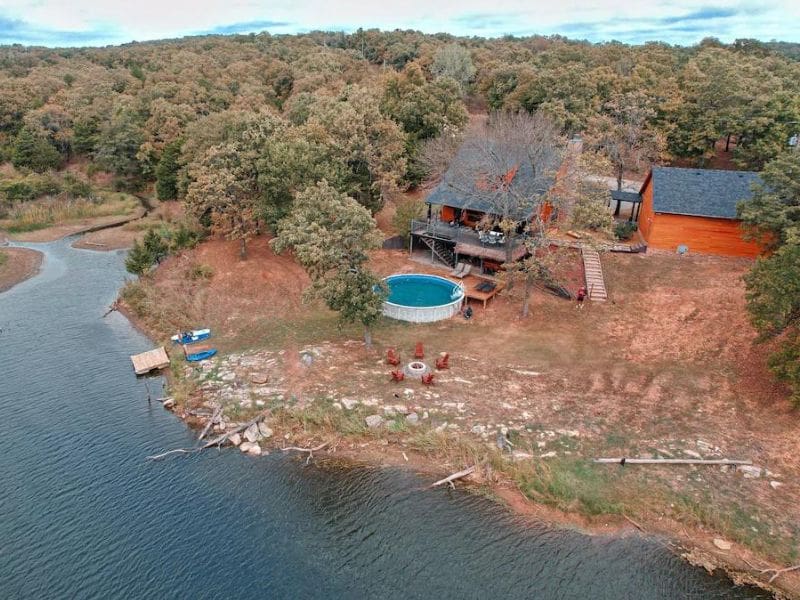 Aerial view of EdgeWater Escape cabin near Turner Falls, featuring a deck, a pool, a fire pit area with chairs, and a lake with surrounding trees.