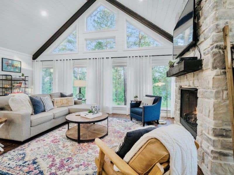 Cozy living room corner with a stone fireplace, comfortable seating, and large windows flooding the space with natural light in a Broken Bow cabin rental.