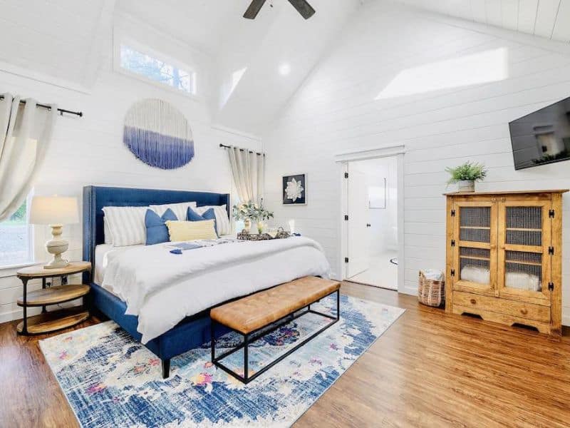 Bright and airy bedroom in a Broken Bow luxury cabin rental featuring a plush blue bed, rustic wooden furniture, and a vibrant area rug on hardwood flooring.