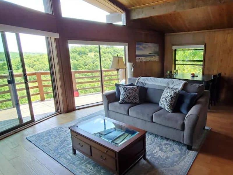 Living room in Lakeview Hideaway cabin near Turner Falls with a gray couch, coffee table, large windows, and sliding glass doors leading to a deck with a view of the forest.